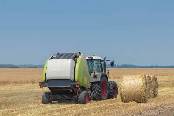 A rear view of a green farming tractor towing a green and white round hay baler in a field next to round hay bales.