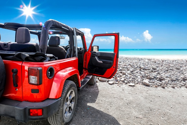 A rear view of a red, two-door Jeep with the top down and driver-side door open, facing a rocky beach on a sunny day.