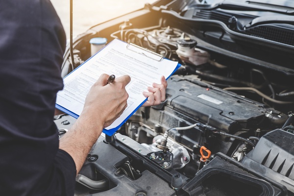 A mechanic with a clipboard and pen filling out a checklist in front of a car with the hood up, showing the engine.