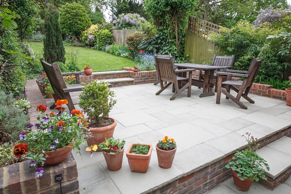 A landscape view of the backyard of a home with a stone and brick patio with wooden furniture and backyard beyond it.