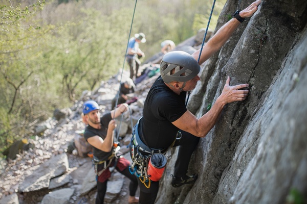 A group of people are standing next to a rock face. One man is in a harness and is climbing up the cliff.