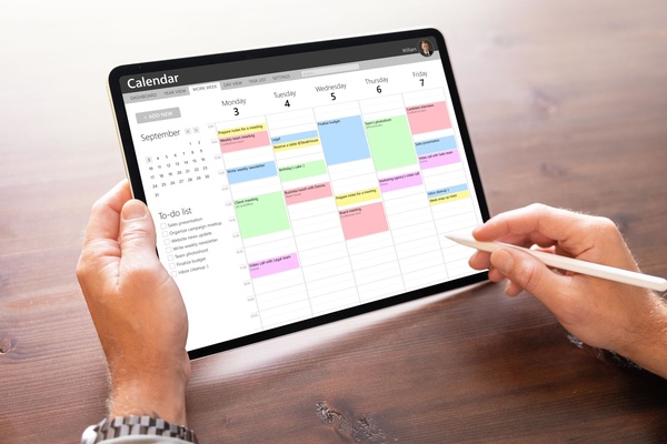 Someone holds a tablet and a white stylus. A calendar is open on the tablet showing a to-do list and a week in September. community bulletin