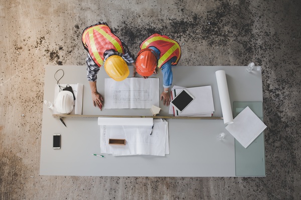 An overhead view of two workers in hard hats and high-visibility vests examining blueprints and papers on a table.
