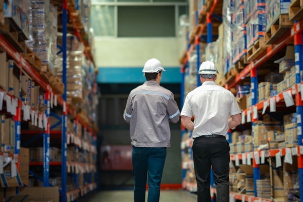 Two employees walking through a row of industrial shelves in a warehouse. They are both wearing hard hats.