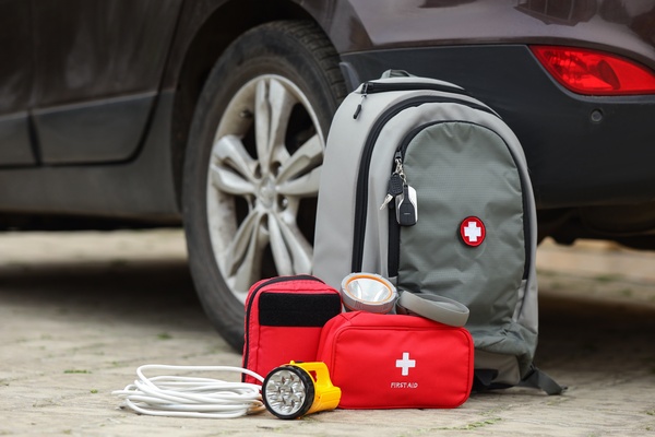 An emergency backpack and first aid kit sitting on the ground next to a car. The backpack has a hospital symbol.