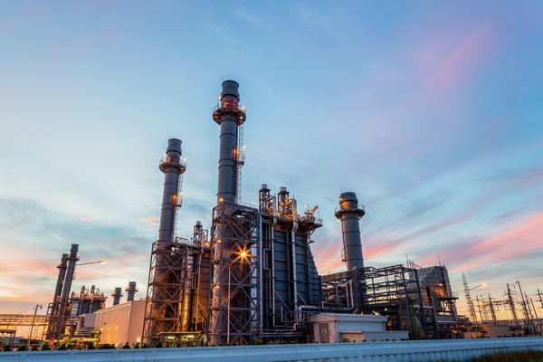 Industrial refinery complex with tall, illuminated smokestacks, metal structures, and pipework against a twilight sky.