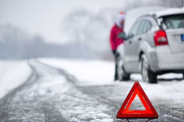 A woman standing beside a car with an open hood on a snowy road with a warning triangle placed behind the vehicle.