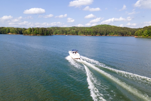 A white pontoon boat cruising across a lake surface and leaving white waves in its wake. The surrounding trees are green.
