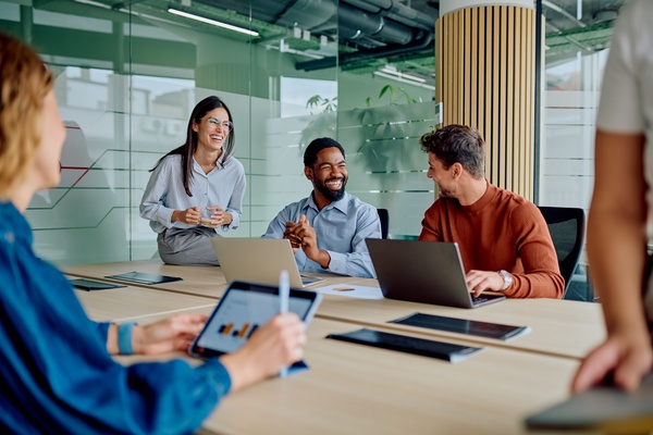 A team of five individuals sits in a modern office, smiling and sharing ideas around a table with laptops.