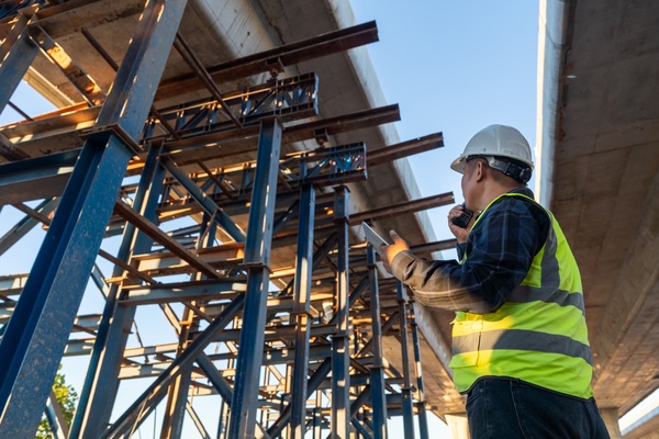 A person in a safety vest and hard hat standing on a road construction site, holding papers and looking over the area.