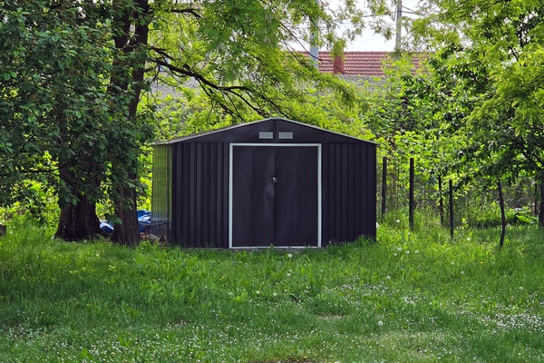 A metal shed sits in a backyard with its doors shut. It stands next to a tree and a garden's wire fence.