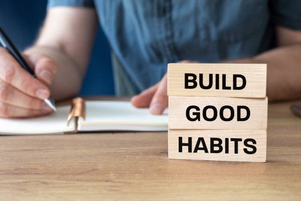 A person sits at a desk, writing in a notebook. Three wooden blocks sit in front of them with the words "Build Good Habits."