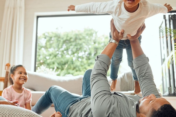 A man is lying on his back as he holds his son above him. A woman is sitting next to them and another child is in the background.
