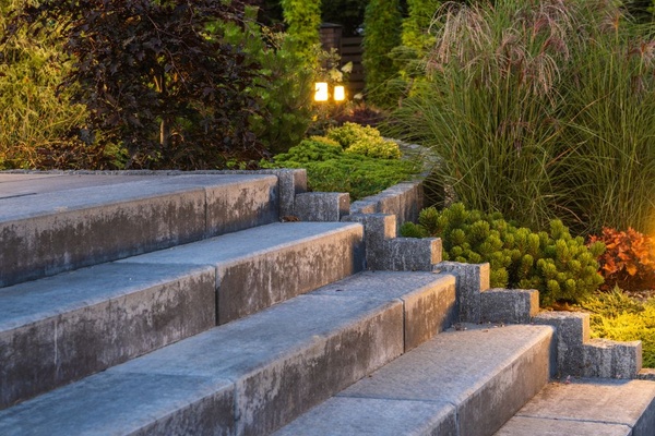 Tall, skinny stake lights illuminate the concrete steps, flowers, and bushes in a residential backyard.
