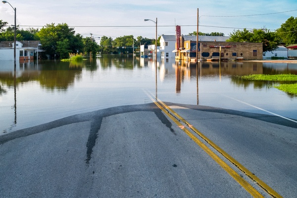 A flooded roadway extending toward partially submerged storefronts, utility poles, trees, and reflected buildings.