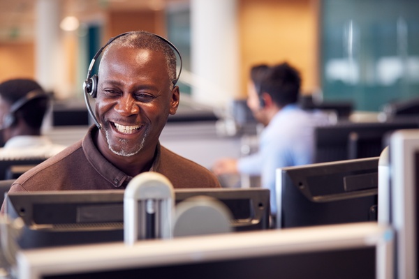 A man wearing a telephone headset and a brown button-up shirt sits at a desk in an office workspace and smiles brightly.