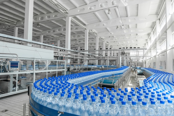 A large industrial facility with a wide conveyor belt moving thousands of clear plastic bottles with blue caps.