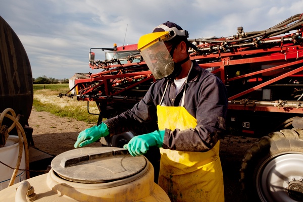 A farmer in chemical safety gear putting the lid back onto a chemical storage container with a large vehicle behind him.
