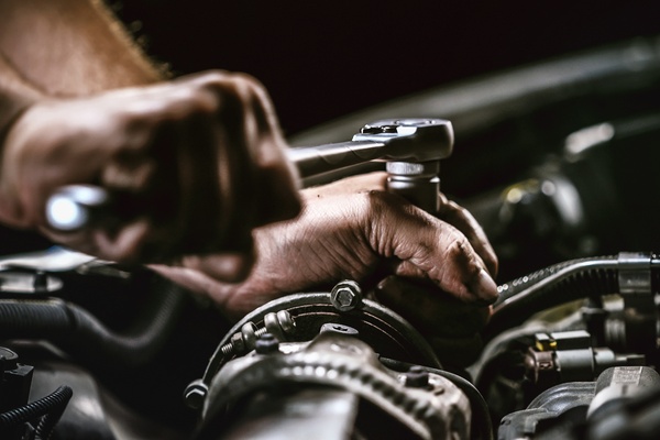 A close-up of a pair of hands using a socket wrench to rotate a nut on a tube inside the engine of a car.