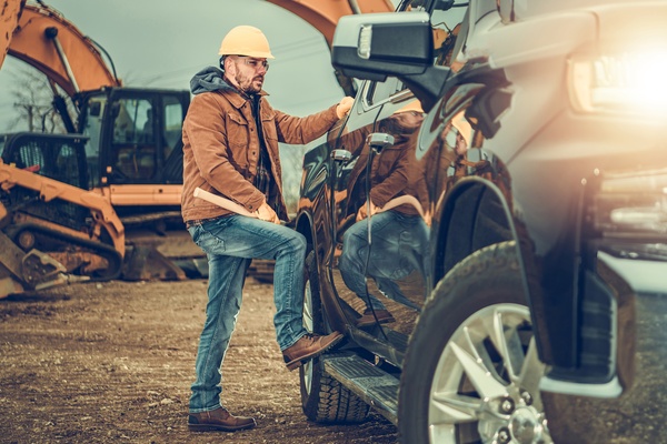 A man wearing a yellow helmet, with a foot on the stepping bar of a black pickup truck on a construction site.