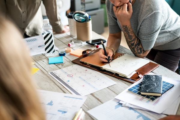  Team members review a calendar and charts while one person takes notes in an open planner at a shared table.