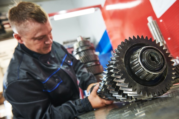 A close-up view shows a male technician touching and inspecting an industrial gearbox. The background is blurry.