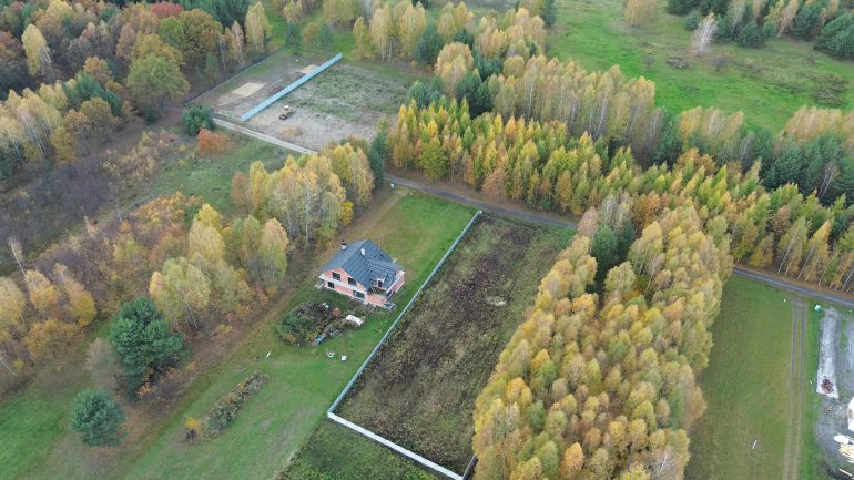 An aerial view of a single small house built in the middle of a field. The field is surrounded by trees.
