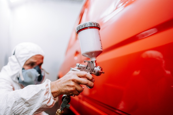 A technician in white protective gear uses a paint spray gun to apply red coating to a vehicle panel.