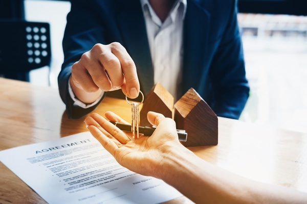 A close-up of the hand of a man holding a set of keys over another hand on top of a desk with an “agreement” document.