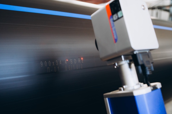 A close-up of a laser marking machine marking letters and numbers on a large black plastic water pipe.