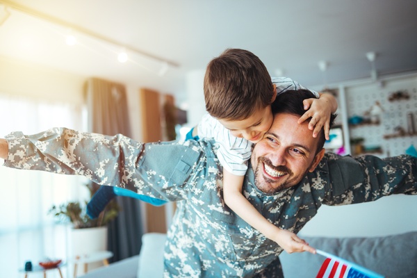 A young boy is sitting on the back of his dad. The dad is leaning over while putting his arms out like an airplane.