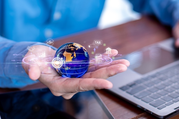 A man's hand holds a globe graphic with business and financial icons displayed as he sits at a desk with a laptop.