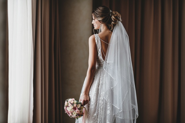 A bride wearing a lace dress and long veil stands beside curtains while holding a small bouquet of pale flowers.