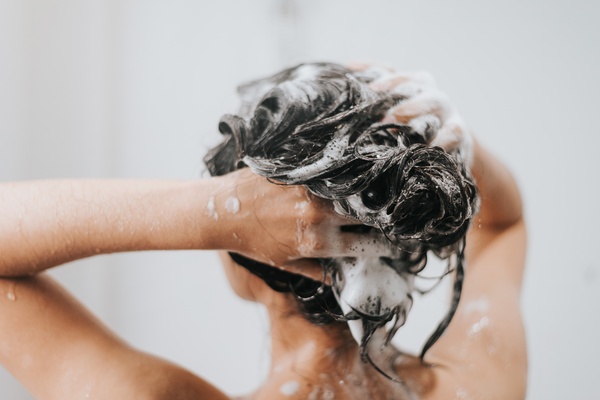 The back of someone's head in the shower. They scrub their scalp with soap, with suds all over their hands and hair.