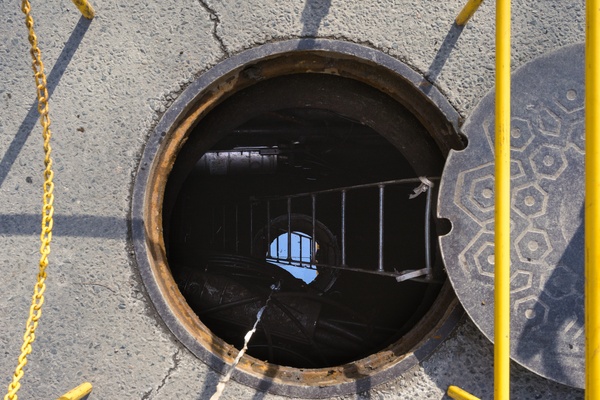 An open manhole with a metal ladder, yellow barrier poles, and a round cover on a cracked concrete street.