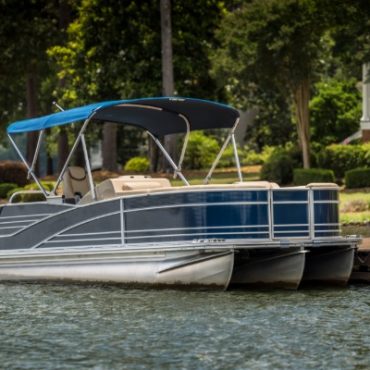Dark blue pontoon boat with a canopy docked on a calm lake beside a tree-lined residential waterfront.
