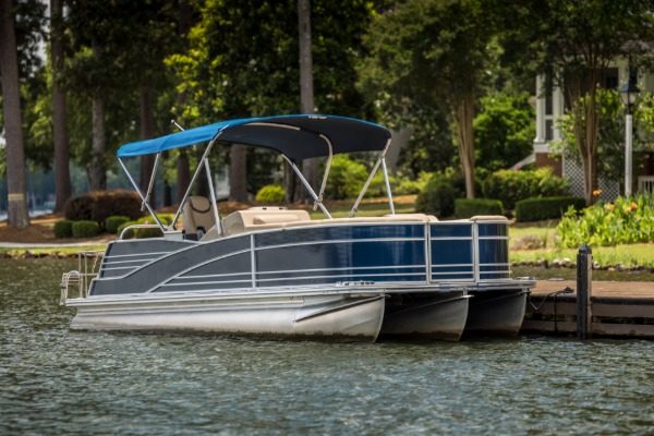Dark blue pontoon boat with a canopy docked on a calm lake beside a tree-lined residential waterfront.