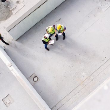 Three people are standing next to each other in a room under construction. There is a fourth person off to the side.