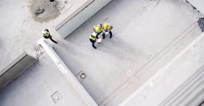 Three people are standing next to each other in a room under construction. There is a fourth person off to the side.