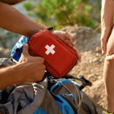 A close-up of a man holding a small first aid kit next to a backpack on a trail outside, as a woman holds her knee in pain.