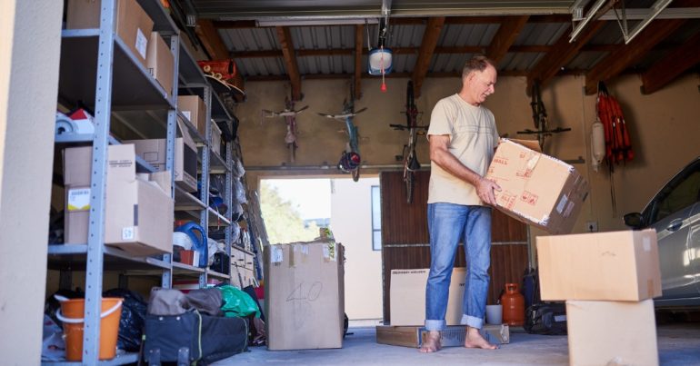 A man without shoes is standing in the middle of a garage with the large door open. He is holding a cardboard box.