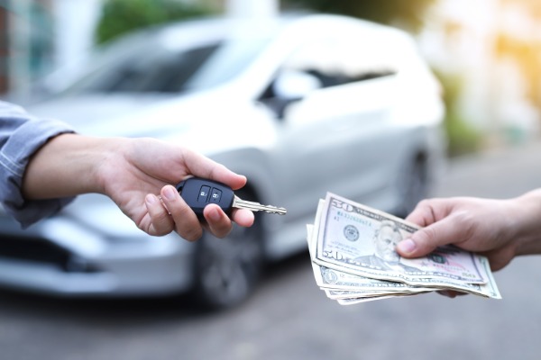 Two hands exchanging car keys and a stack of cash in front of a blurred white car parked on a street.