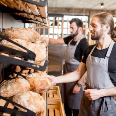 Two men in black T-shirts with grey aprons and clear plastic gloves grabbing bread from the shelves of a bakery.
