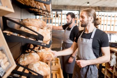 Two men in black T-shirts with grey aprons and clear plastic gloves grabbing bread from the shelves of a bakery.