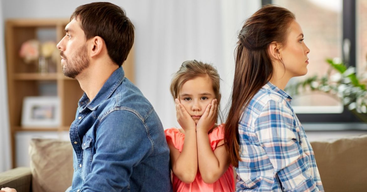 A little girl holding her face with both hands, sitting between a woman and a man looking in opposite directions.