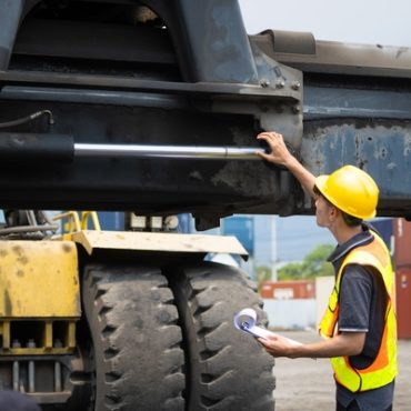 A worker in a yellow hard hat and safety vest inspecting a large crane truck while holding a clipboard.