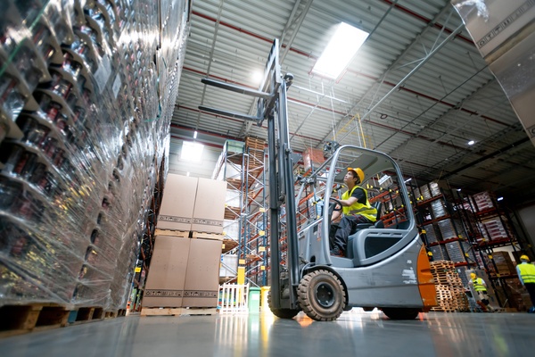 A warehouse worker operates a forklift beside stacked pallets. Tall racks and more workers are in the background.
