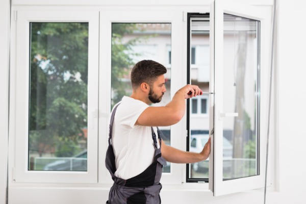 A worker using a screwdriver on an open white window frame, with large glass panes and greenery outside.
