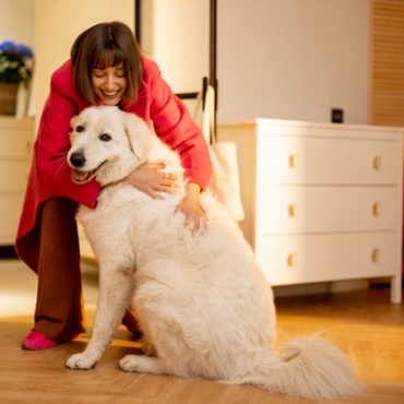 A smiling woman in a red coat hugs a large white dog inside a warmly lit bedroom near an open doorway.