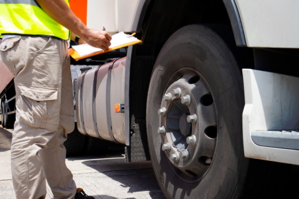 A safety worker writes on a clipboard while inspecting the front wheel and fuel tank of a commercial truck.